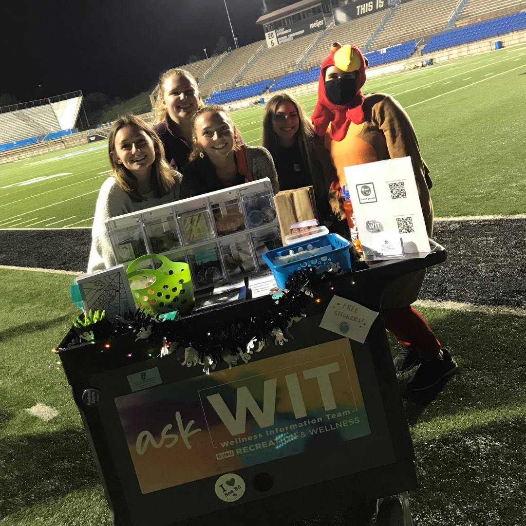 Image of five RecWell staff behind the Ask WIT cart in Lubbers stadium. One is wearing a turkey costume.